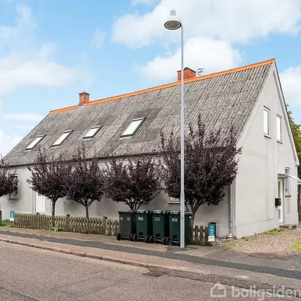 Et gråt hus med skråt tag står ved en gade; træer er plantet foran et hegn. En lygtepæl ses i forgrunden, og en garage er i baggrunden.