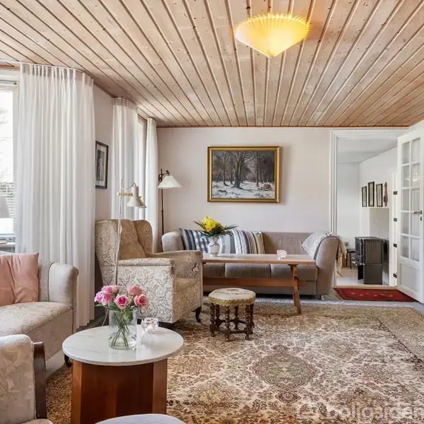 Stue med to bløde sofaer og et sofabord, dekoreret med blomster og puder. Rummet har tæppe, træloft, vægmaleri, samt rigeligt med naturligt lys fra vinduerne.