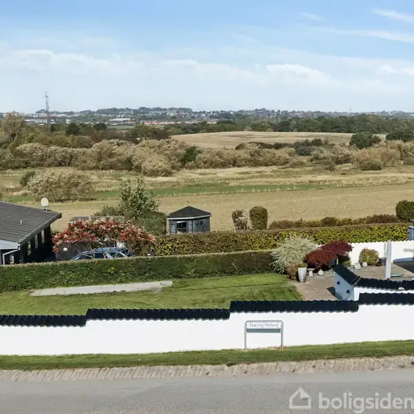 Hus med hvid facade og sort tag omkranset af hæk, beliggende i et landligt område med marker i baggrunden. Grøn græsplæne og indkørsel foran huset. Skilt: "Fællesantenne".