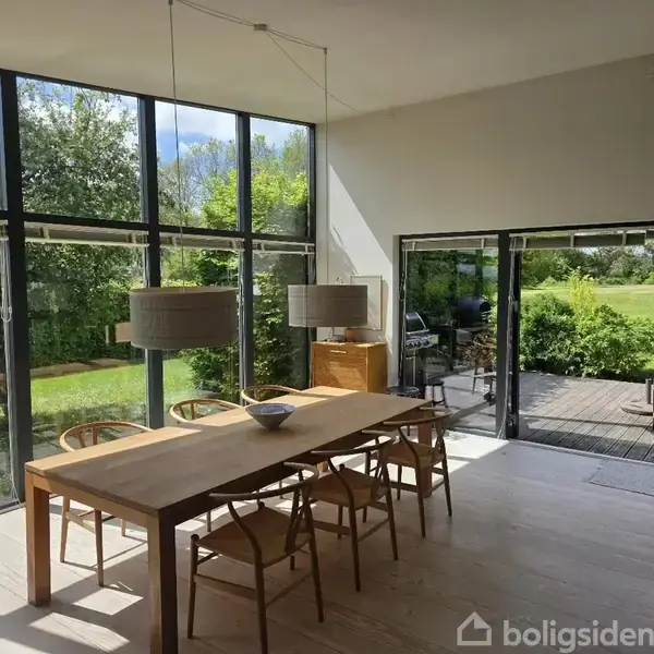 Et spisebord står i en lys stue med store vinduer, der åbner op til en grøn have og terrasse. To lamper hænger over bordet, omgivet af træmøbler.