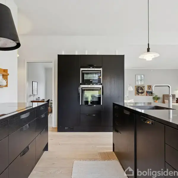 Et moderne køkken med sorte skabe og indbyggede ovne. To hængende lamper over køkkenøen. I baggrunden ses en lys stue med bøger og billeder.