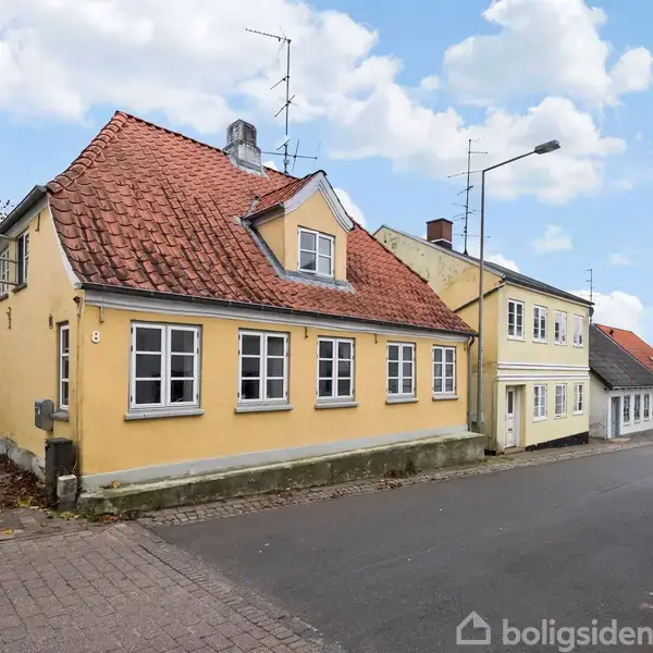 Et gult hus med rødt tegltag står ved en stille gade med brosten og få træer i baggrunden. Husets vinduer har hvide sprosser. Nummer "8" ses ved indgangsdøren.