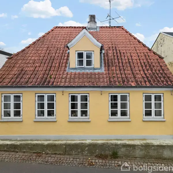 Gult hus med rødt tegltag og fem hvide vinduer. Det står i en bymæssig gade med brosten og tilstødende bygninger.