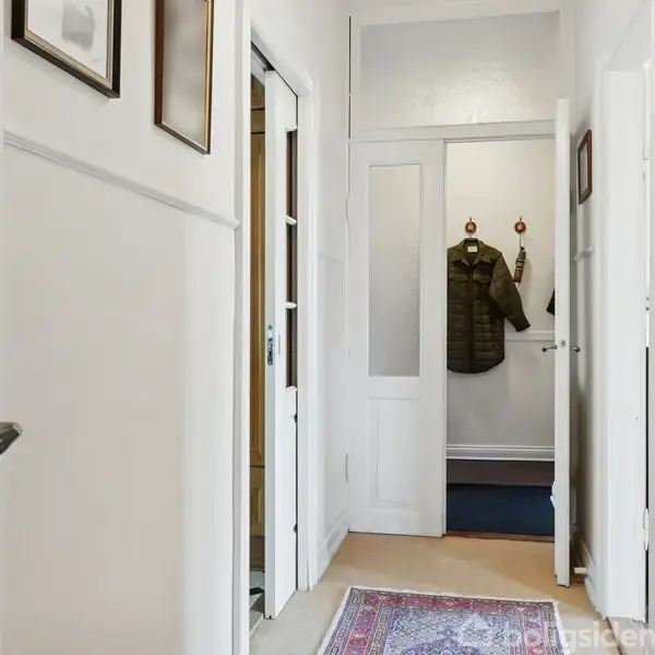Gang med et tæppe på gulvet og indrammede billeder på væggene. Jakker hænger på knager i baggrunden nær en hvid dør, der fører til et andet rum.