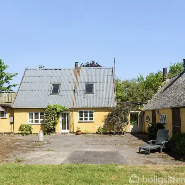 Gult hus med skråtag omgivet af gårdsplads, omkranset af to mindre bygninger med stråtag. Græsplæne i forgrunden, grønne buske og træer i baggrunden under blå himmel.