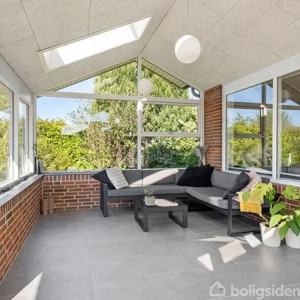 Grå hjørnesofa står i en solrig vinterhave med store vinduer, murstensvægge og loftsvinduer. To bølgeplanter og små puder dekorerer rummet, med en udendørs udsigt af grønne buske.