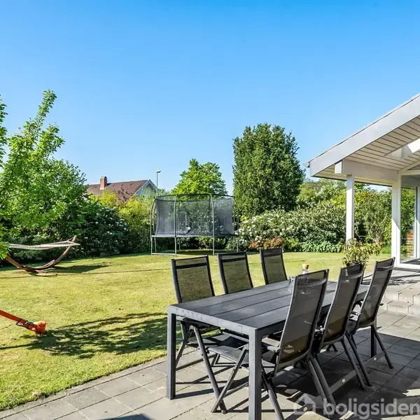 Et sort udendørs spisebord med otte stole står på en solrig terrasse. Ved siden af er en græsplæne med et hængekøjestativ og en trampolin nær en moderne tilbygning.