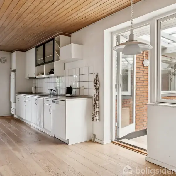 Køkken med hvide skabe står i et rum med trægulv og træloft. En glasdør åbner mod en indendørs terrasse med røde murstensvægge. Lampe hænger fra loftet.