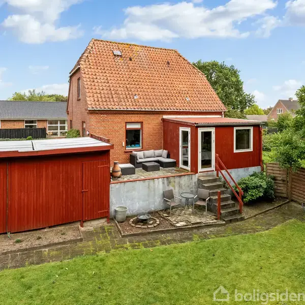 Rødt murstenshus med rødt tegltag står stille. Haven rummer en terrasse med havemøbler, omgivet af grøn græsplæne og hegn. Et skur læner sig op ad huset.