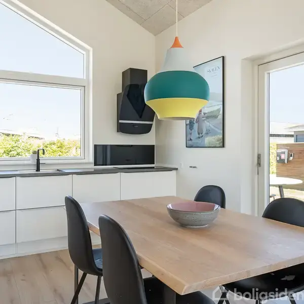 Bord med fire stole står under en farverig lampe i et lyst køkken med minimalistiske hvide skabe. Billedet viser også en glasdør og store vinduer ud til en have.