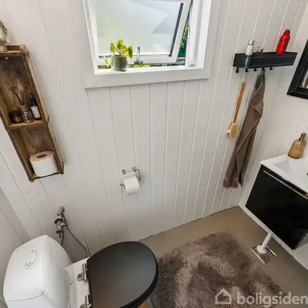 Toilet med sort sæde, placeret i lille badeværelse med hvidmalede vægge. Over toilettet er der en træhylde med en Buddha-figur. Håndvask med sort skab og spejl findes til højre. En lille plante står i vinduet.