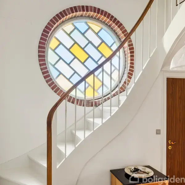 En spiraltrappe snor sig opad ved siden af et rundt vindue med farvede ruder i et rum med hvidmalede vægge og moderne kunst på væggen.