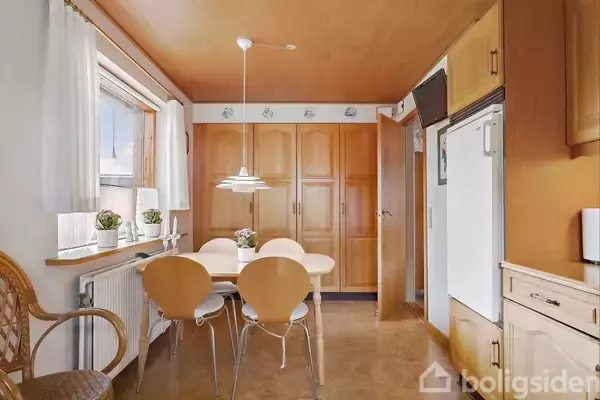 Et lyst køkken med et rundt spisebord og fire stole, omgivet af træskabe og store vinduer med blomster. En loftslampe hænger over bordet.