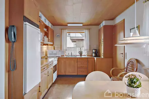 Køkken i træ med køleskab, komfur og mikrobølgeovn langs væg. Rundt bord med stole i forgrunden. Rummets lysebrune farvetone skaber en hyggelig atmosfære.