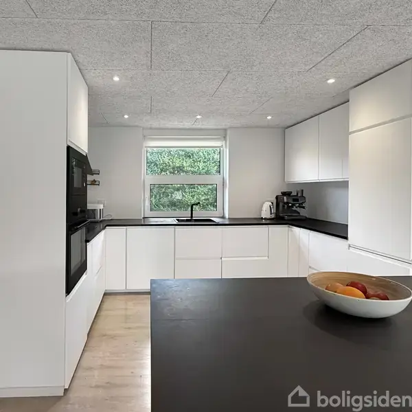 Et moderne køkken med hvidt køkkeninventar og sort bordplade. En skål med frugt står på bordet. Store vinduer i baggrunden giver udsyn til en grøn have.