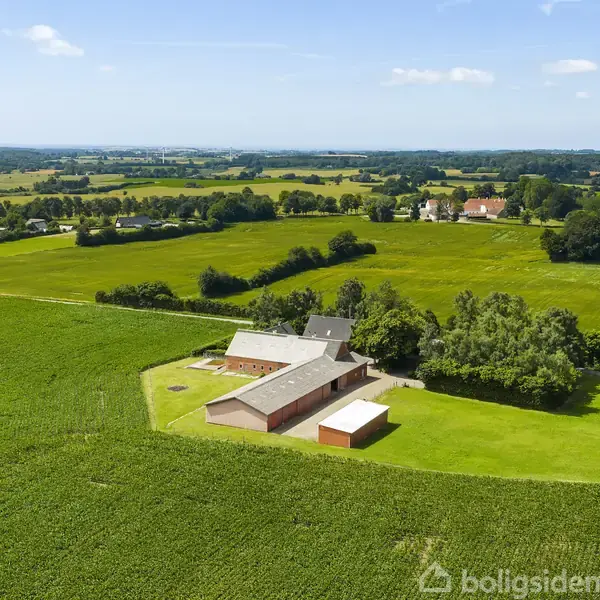 En gård med flere bygninger er omgivet af store, grønne marker og træer. Bygningerne ligger centralt i billedet, mens det åbne landskab strækker sig mod horisonten under en klar himmel.