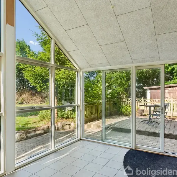 En glaspavillon med skråt tag står tom, omringet af en flisebelagt terrasse og frodig have. Udendørs ses en terrasse med træmøbler og grønne træer i baggrunden.
