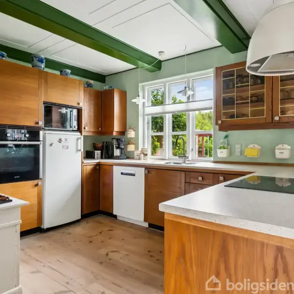 Køkken med træskabe og grønne bjælker indeholder en ovn, mikrobølgeovn og opvaskemaskine. Store vinduer giver udsigt til natur. En hvid loftslampe hænger over en lys køkkenø.