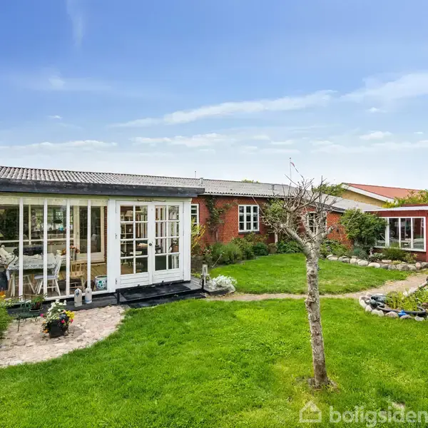 Hus med store vinduer står solbelyst i en grøn have. Terrassen har blomsterkrukker, og græsplænen omkranses af beplantning og en rød træbygning i baggrunden.