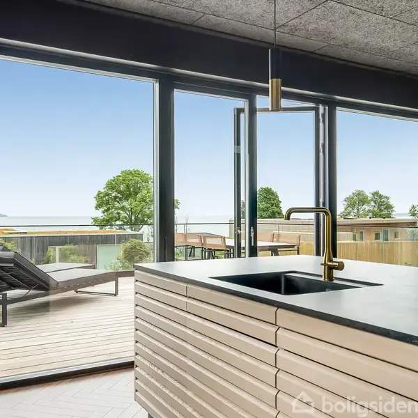 Køkkenø i moderne stil med håndvask og guldhane foran store glasdøre. Dørene åbner mod en terrasse med liggestole, og udsigt til træer og havet i baggrunden.