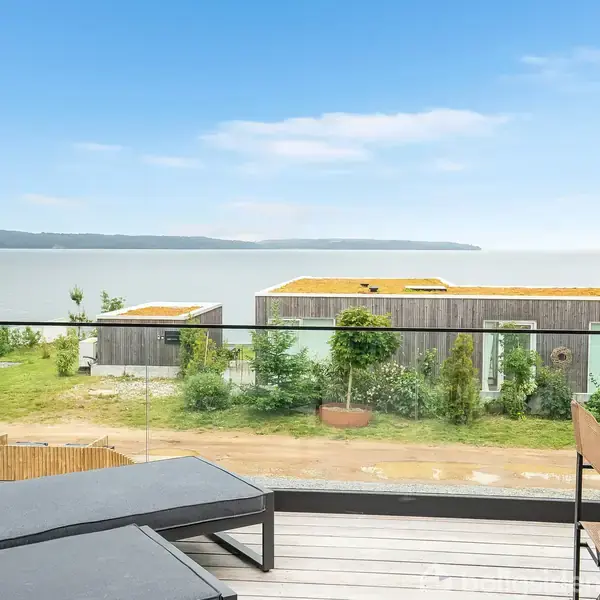 Liggestole står på en terrasse med udsigt over vandet og moderne bygninger med grønne tage. Træer og græs omkranser bygningerne, under en klar blå himmel.
