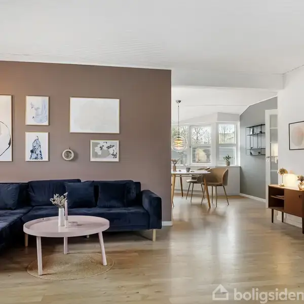 En blå sofa står ved en brun væg med kunst, ved siden af et rundt bord. Lyse trægulve omgiver rummet, som åbner op mod en spisestue med vinduer.