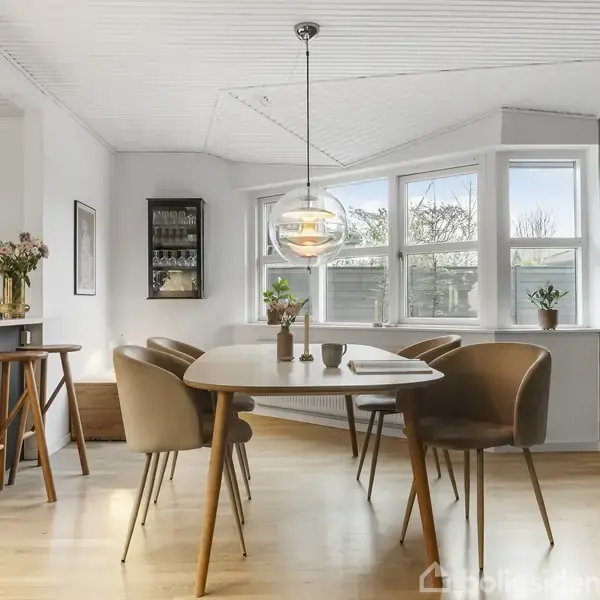 Et spisebord med seks brune stole står i et lyst rum. En pendellampe hænger over bordet. Der er store vinduer i baggrunden og en køkkenø med blomster til venstre.