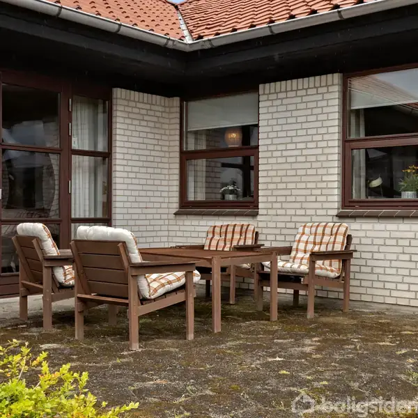 Terrassemøbler står stille på en stenbelagt terrasse foran et hvidt murstenshus med store vinduer og en dør. Stole har hynder, og der er buske i forgrunden.