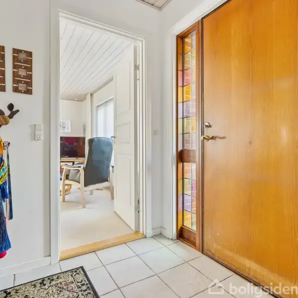 En entré med trædør med farvede glasfelter, åbner mod et kontor med lænestol. Dekorationer og jakker hænger på væggen ved siden af et lille bord på flisegulv.