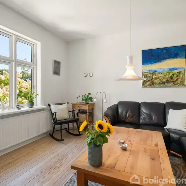 Sort lædersofa står ved siden af et lyst træbord i en stue. Solskin kommer ind gennem store vinduer med planter på karmen. En gyngestol og skrivebord ses i hjørnet. Væggen har et maleri og en hængende lampe.