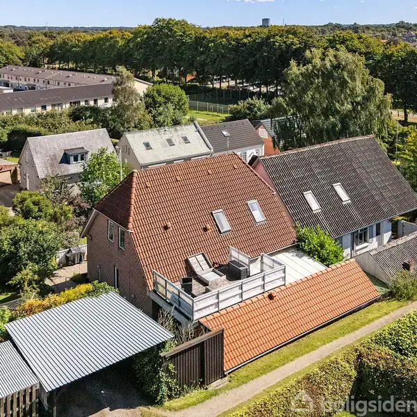 Et hus med en stor terrasse på taget ses i en række af boliger omgivet af træer og haver i et villakvarter.