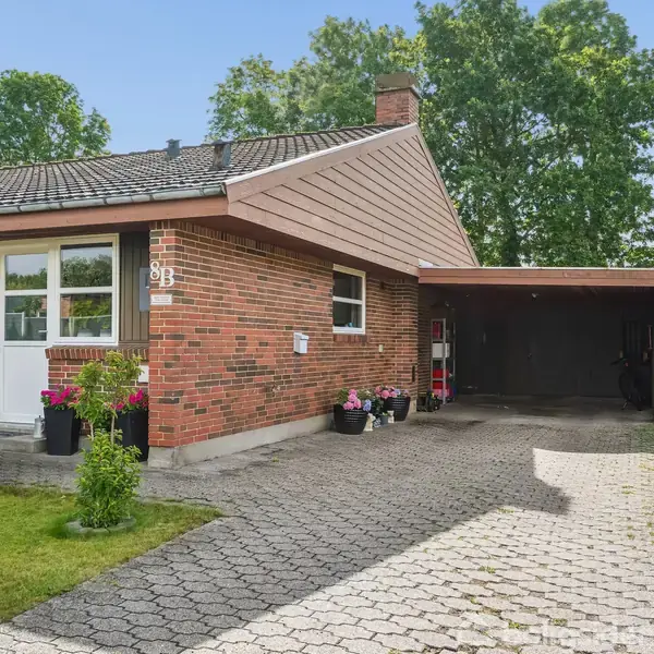 Et rødstenshus med en forlænget tagkonstruktion danner en carport. Huset har vinduer og en dør samt planter foran. Græsområde omkranser indkørslen til carporten. Nummer "9B" vises ved indgangen.