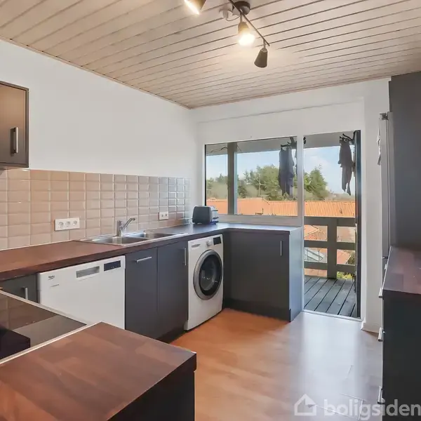 Køkken med brun træbordplade fungerer i moderne stil, indeholder kogeplade, opvaskemaskine og vaskemaskine. Omgivelserne inkluderer lyse vægge, et vindue mod terrasse og træbeklædt loft.