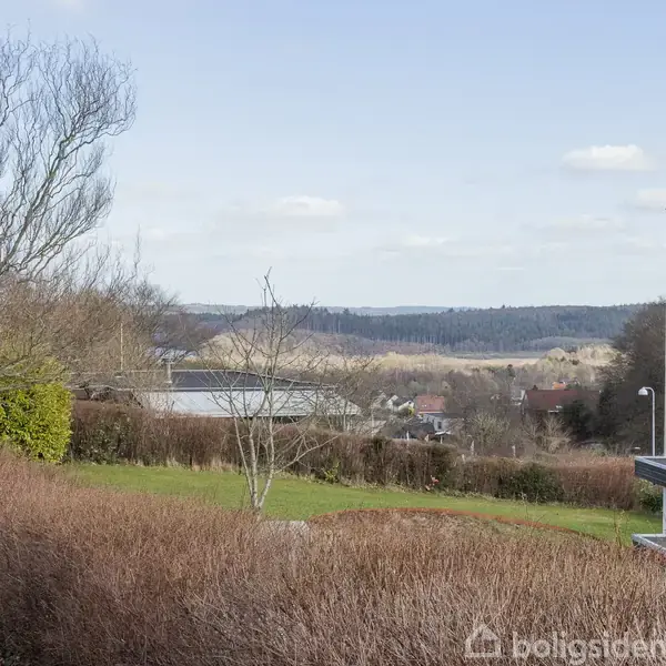 Træer står i en have med udsigt over huse og landskab. Buske afgrænser området, og en bygning ses til højre. Himmelen er klar med få skyer.