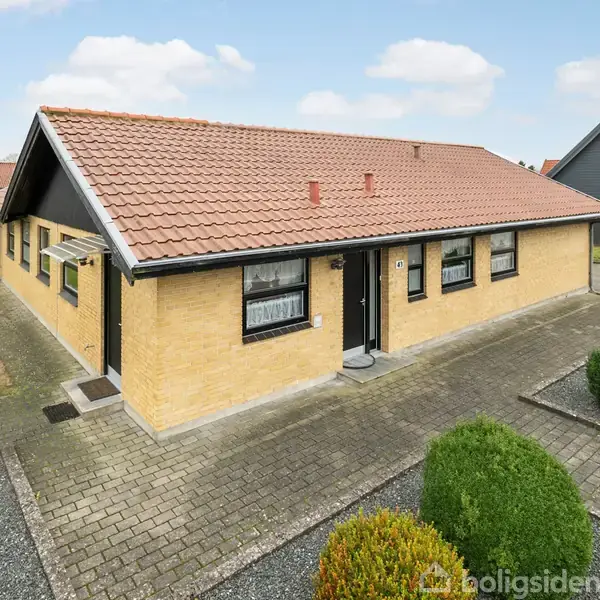 Et murstenshus med rødt tegltag står på en flisebelagt indkørsel. Huset er omgivet af en velholdt have med buske. Himlen er blå med spredte skyer.