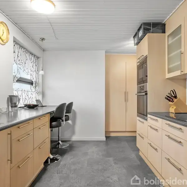 Et køkken med lyse træskabe står i fokus. En integreret ovn og forskellige køkkenredskaber er placeret på bordpladerne. Rummet er lyst med blomsterdekorerede gardiner og en gul vægur.