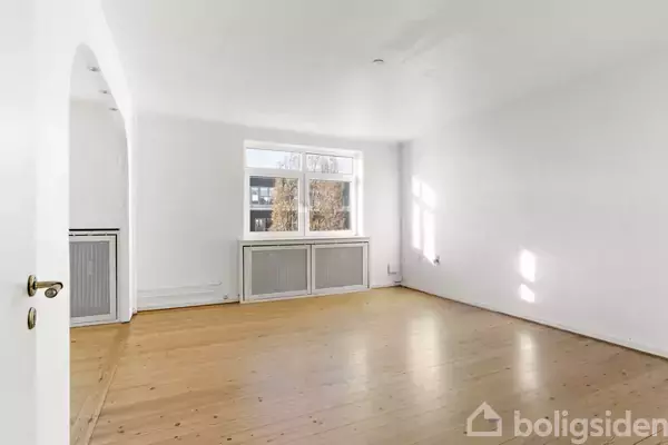 Et tomt rum med trægulv og hvidmalede vægge, oplyst af naturligt lys fra et stort vindue. Til venstre ses en åbning til et tilstødende rum.