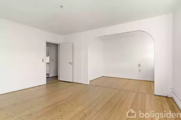 Rummeligt værelse med trægulve og hvide vægge. En dør fører til et tilstødende rum, og en stor bueåbning forbinder to rum i et minimalistisk interiør.