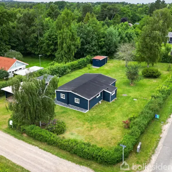 Et blåt hus ligger på en velplejet græsplæne, omgivet af buske og træer. En grusvej ses foran huset, med en skov i baggrunden.