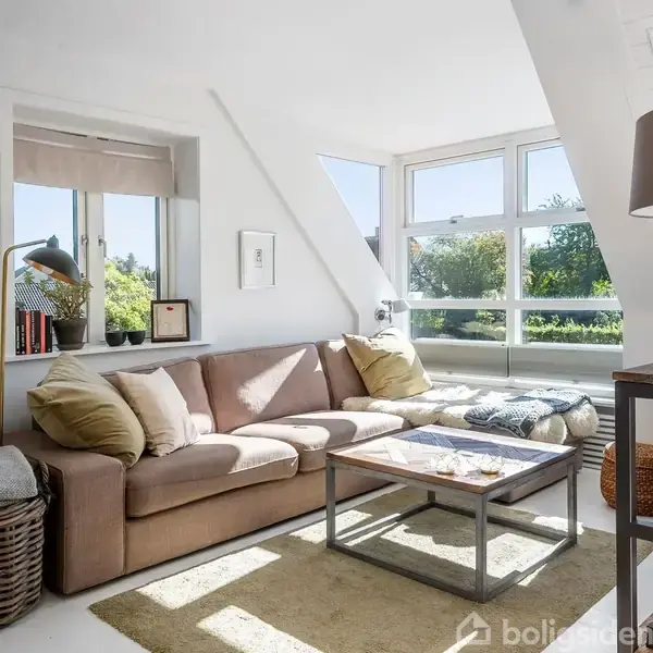 En beige sofa med puder står i en lys stue ved siden af et stort hjørnevindue. Bøger og en lampe pynter vindueskarmen. Udsigt til grønt landskab udenfor.