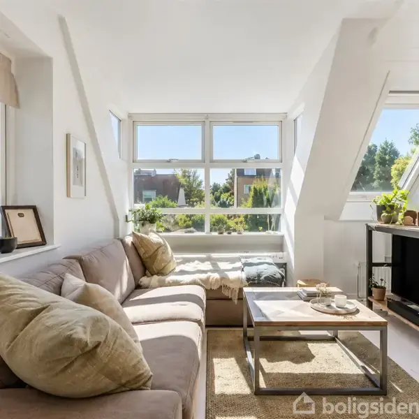 Stor beige sofa fyldt med puder står langs vinduer i en lys stue. Et sofabord med dekorationer står foran, mens en TV-enhed står til højre. Grønne planter pynter rummet.