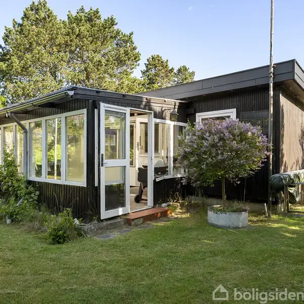 Et lille, sortmalet sommerhus med store vinduer står på en velplejet græsplæne. Huset har en glasindgang og er omgivet af buske og træer under en blå himmel.