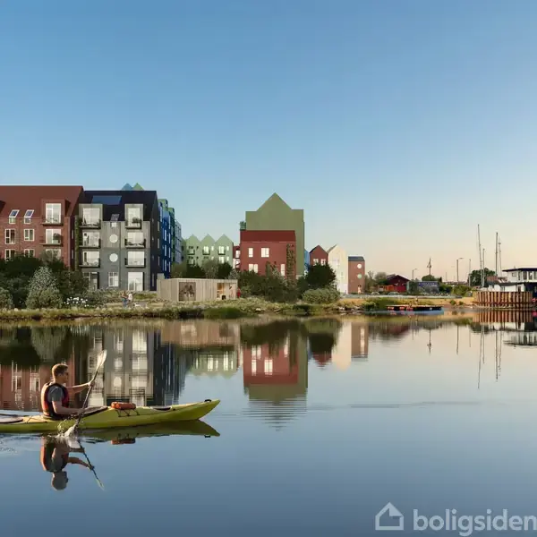 En person ror i en kajak på en stille sø foran en række farverige bygninger. Området omkring søen er bebygget og ligner en urban marina.