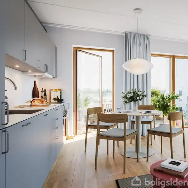 Et lyst køkken med træmøbler viser et bord med blomster og fire stole. Dør fører til balkon med udsigt. Frisk brød på køkkenbordet ved siden af skabsplads.