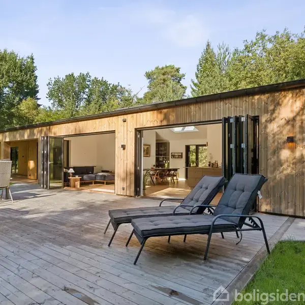 Træhus med store glasdøre åbne mod en terrasse, hvor der står to liggestole og en grill. Huset er omgivet af frodig skov og grønne omgivelser.