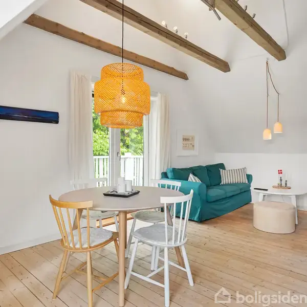 Spisebord med stole står i et lyst rum med skråvægge og synlige træbjælker. Sofa i hjørnet ved vinduet, lysekrone og reoler skaber en hyggelig atmosfære.
