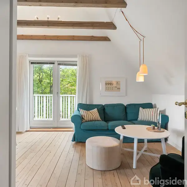 Stue med blå sofa og et rundt bord, belyst af en hængelampe. Store vinduer med udsigt til grønne træer. Trægulve og synlige loftsbjælker skaber en hyggelig atmosfære.