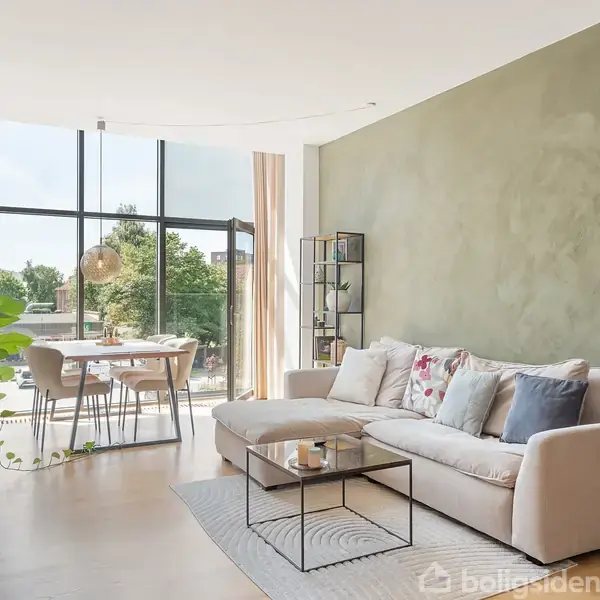 Stue med en hvid sofa fyldt med farverige puder, i en lys, moderne indretning. En stor vinduesvæg i baggrunden lader naturligt lys ind over spisebordet. Grønne planter dekorerer rummet.