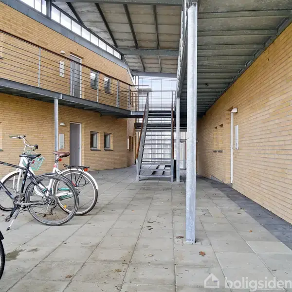 Cykler står parkeret på en flisebelagt gang udenfor en moderne bygning med murstensfacade. Trapper fører til en øverste etage. En dør har tallet "6" ved siden af.