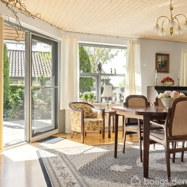 Spisebord med stole står i en stue, badet i sollys fra store vinduer og terrassedør. Rummet har tæppe på trægulv, blomsterdekoration og bløde stole tæt på døren.